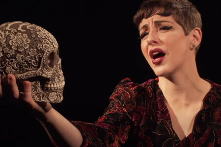 Steffi D. holds a replica of a skull in her right hand. She is singing.