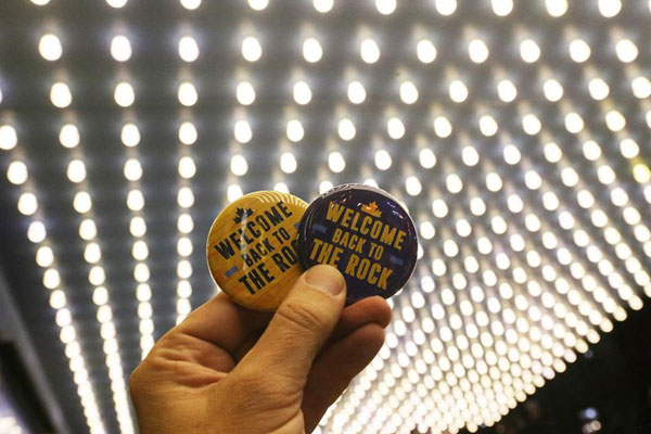Hand holds two show buttons reading welcome back to the rock against a backdrop of the underside of the lighted theatre awning